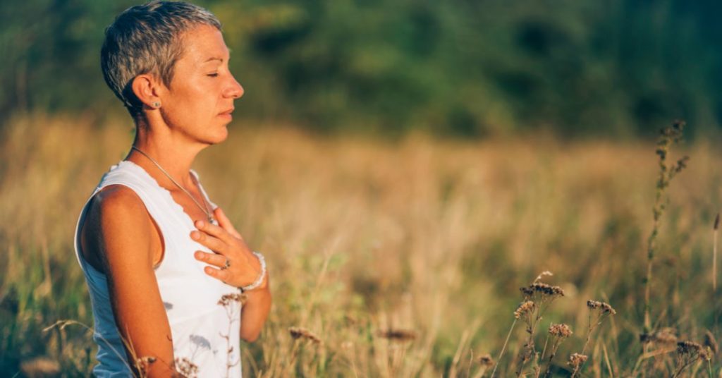 A woman is in a field with her eyes closed and her hand on her heart, soaking up the sun and relaxing, Ways to Stop Sugar Craving
