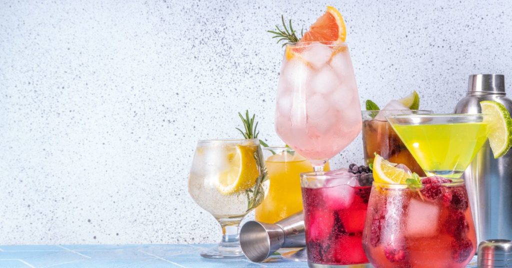 Different types of alcohol glasses and shakers filled with colorful liquids, different fresh fruits and herbs