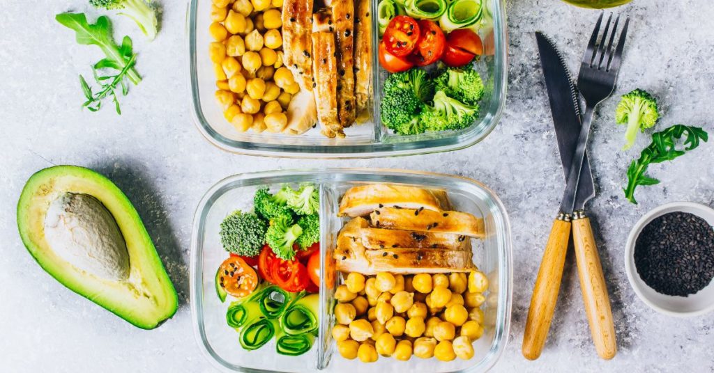 Two glass storage containers filled with prepared foods like chickpeas, raw vegetables, and roast chicken, next to a halved avocado and fork and knife