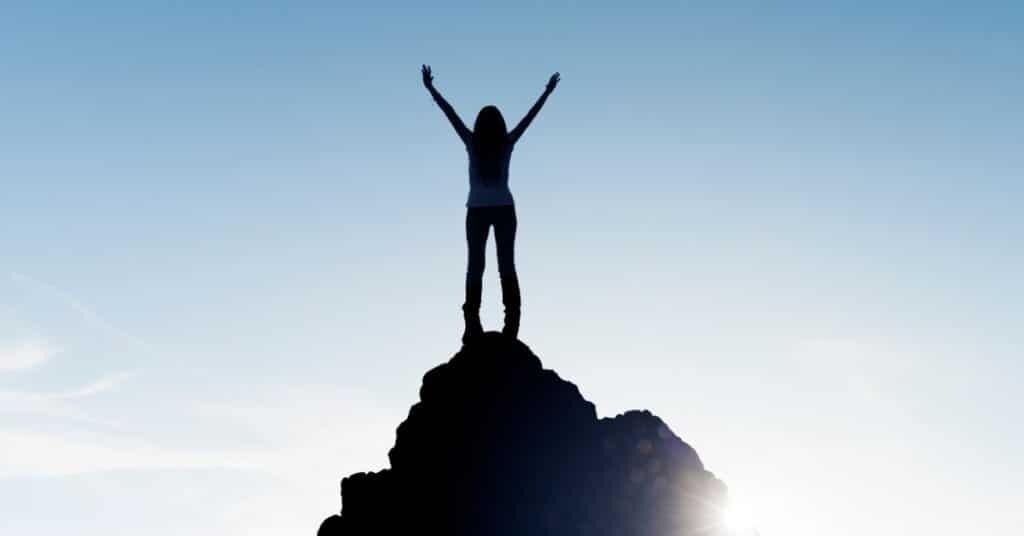 The silhouette of a person stands atop a mountain with arms widespread in celebration of achieving their SMART goal