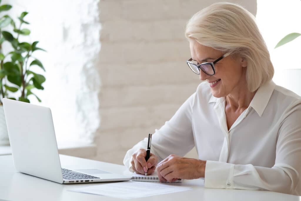 woman sitting at her desk writing goals down in a notebook with a laptop in front of her